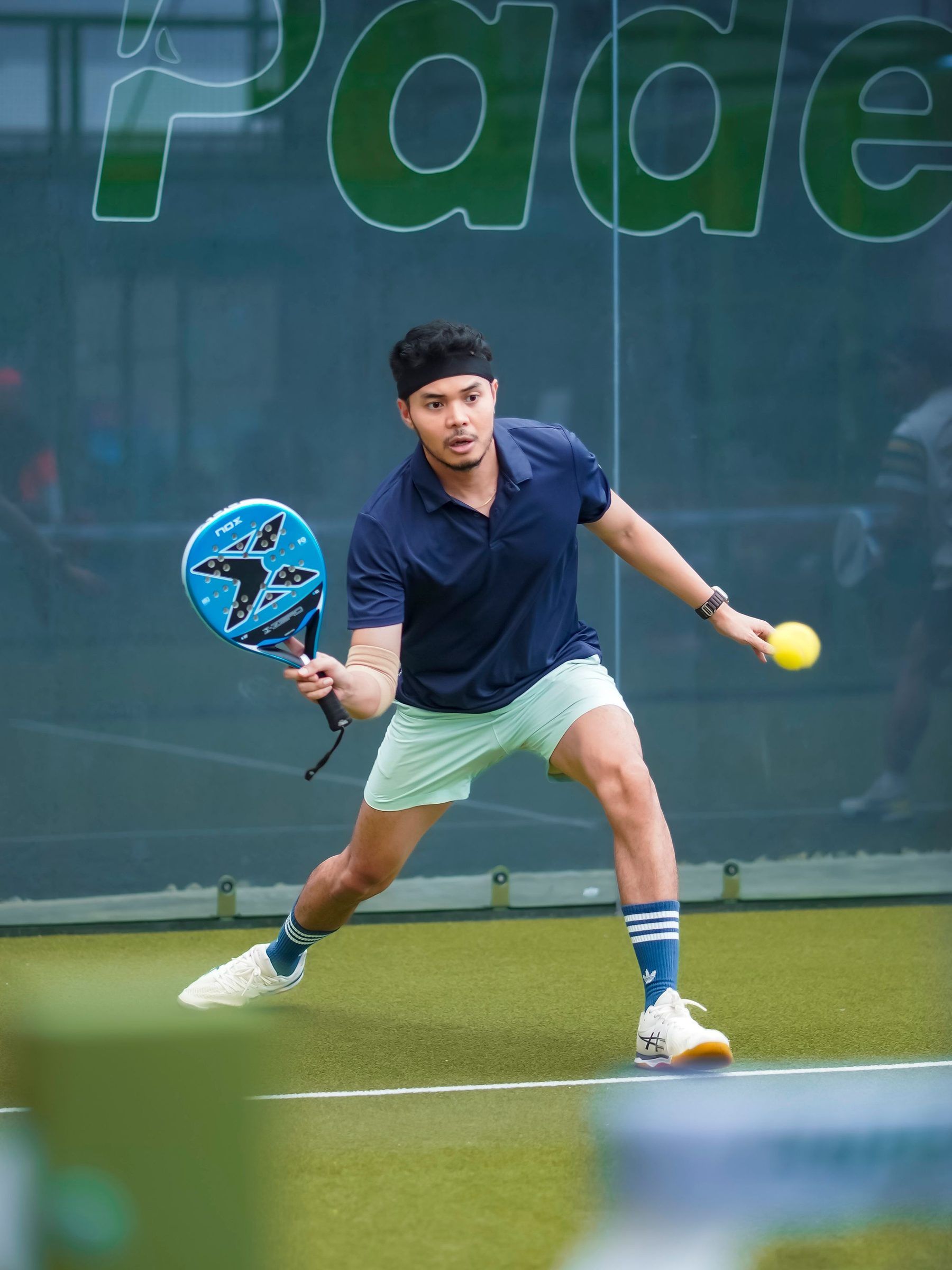 Padelspieler spielt eine einhändige Rückhand auf einem grünen Court vor einer Glaswand