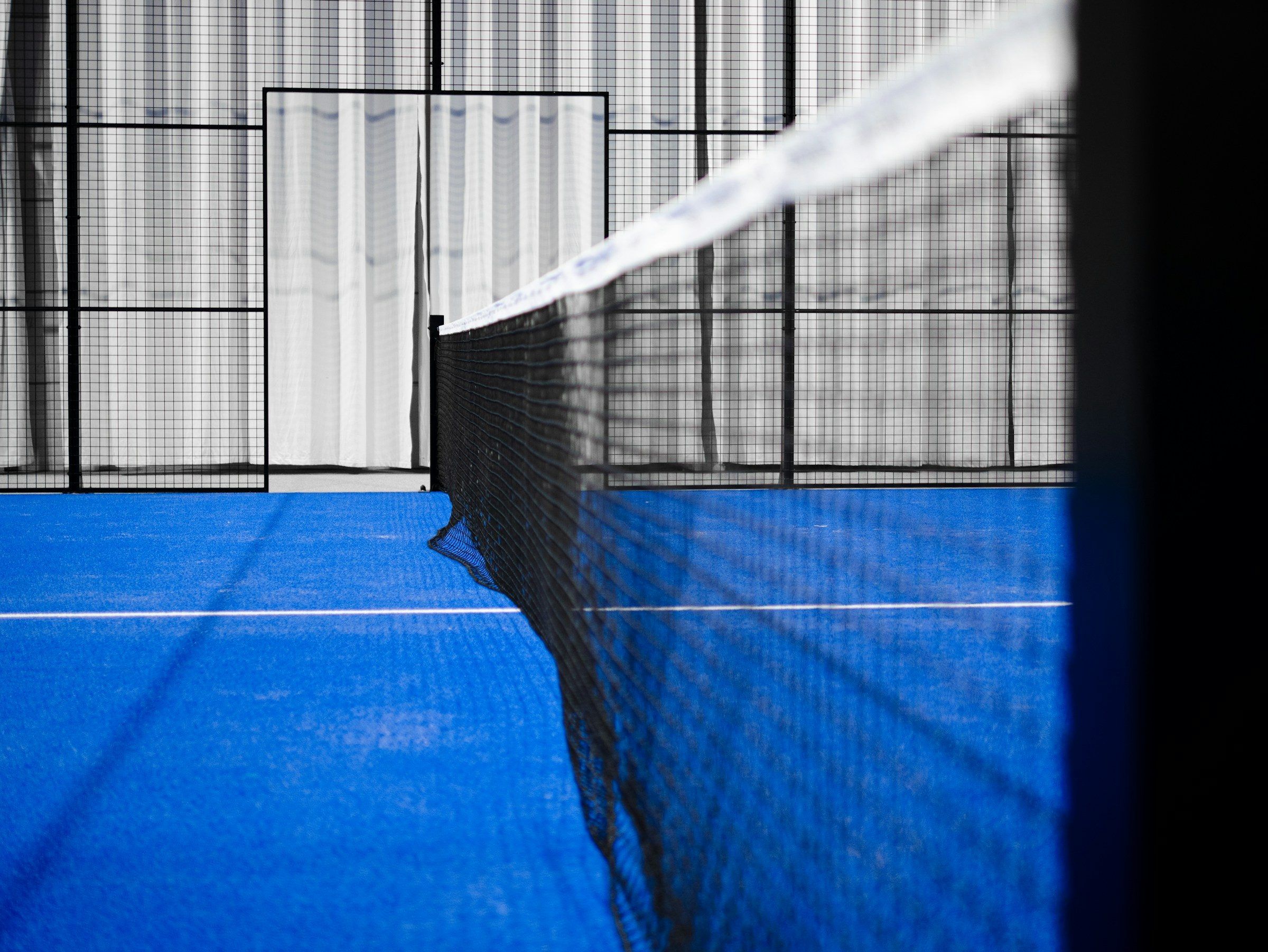 Netz und weiße Linien eines blauen Padelcourts, im Hintergrund Glaswände und Gitter