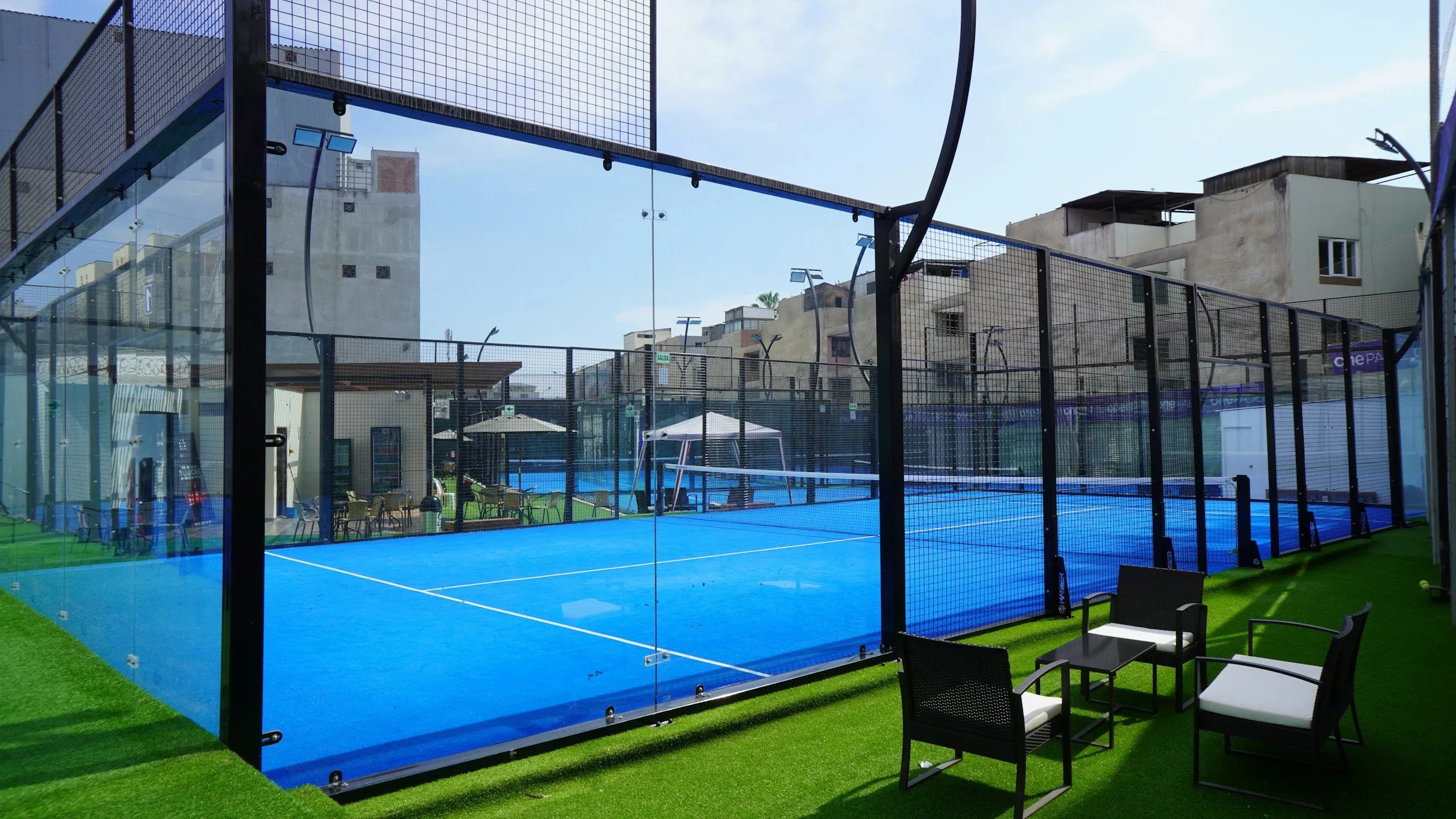 Outdoor-Padelcourt mit Glaswand, daneben Sitzgruppen auf Kunstrasen
