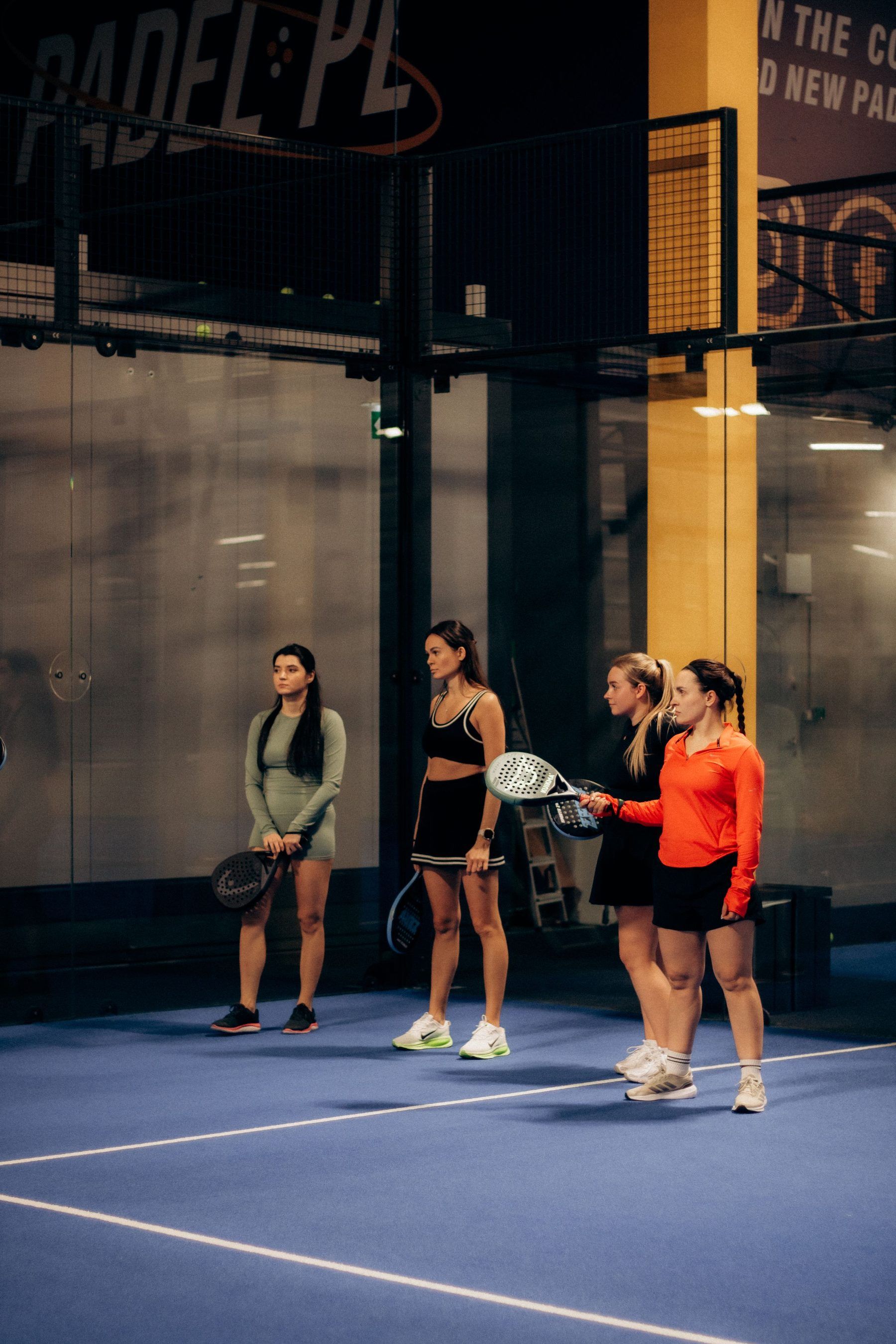 Vier Padelspielerinnen stehen lachend mit ihren Schlägern auf einem blauen Indoor-Court