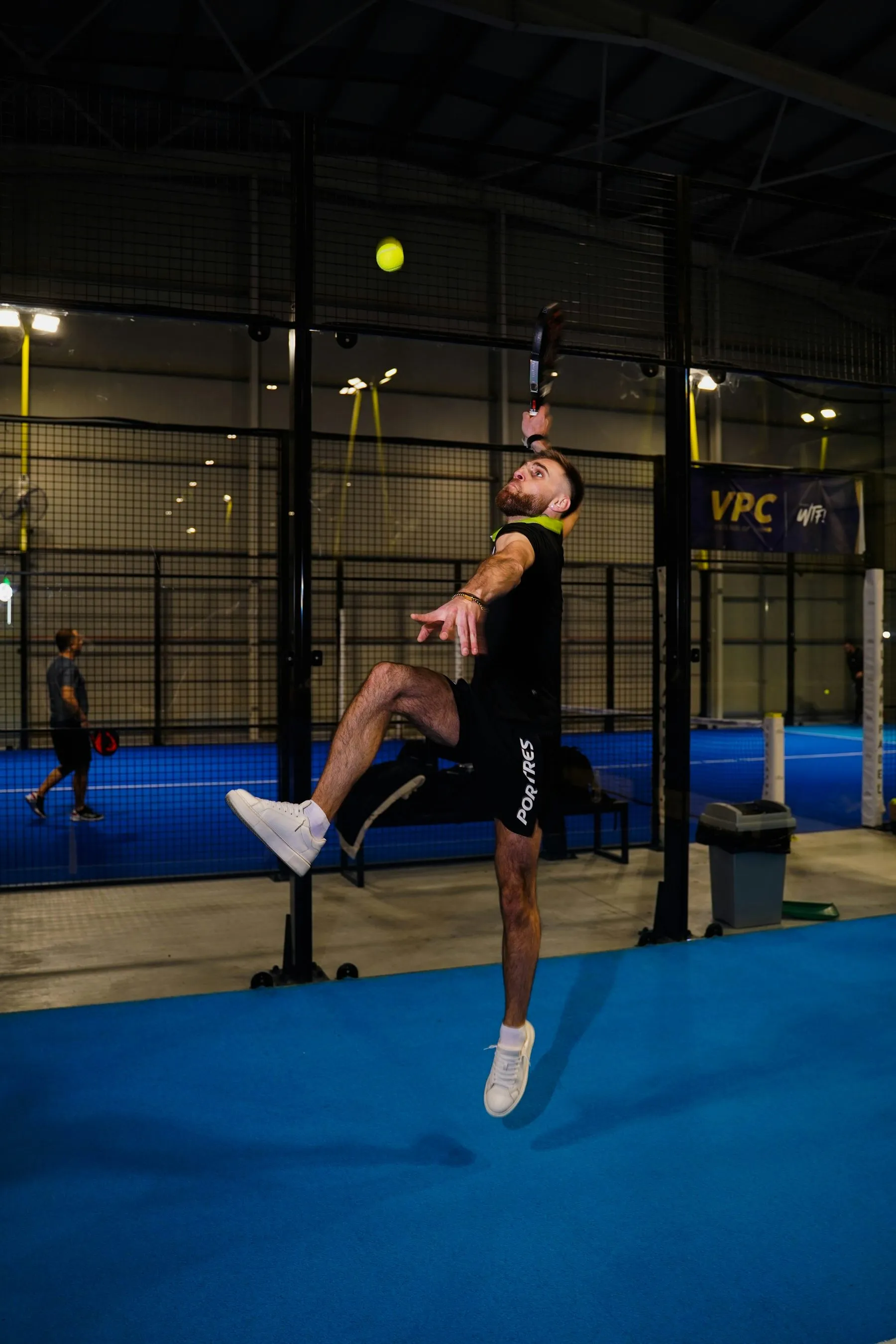 Padelspieler springt in einer Halle zu einem Überkopfschlag, der Ball fliegt über ihm