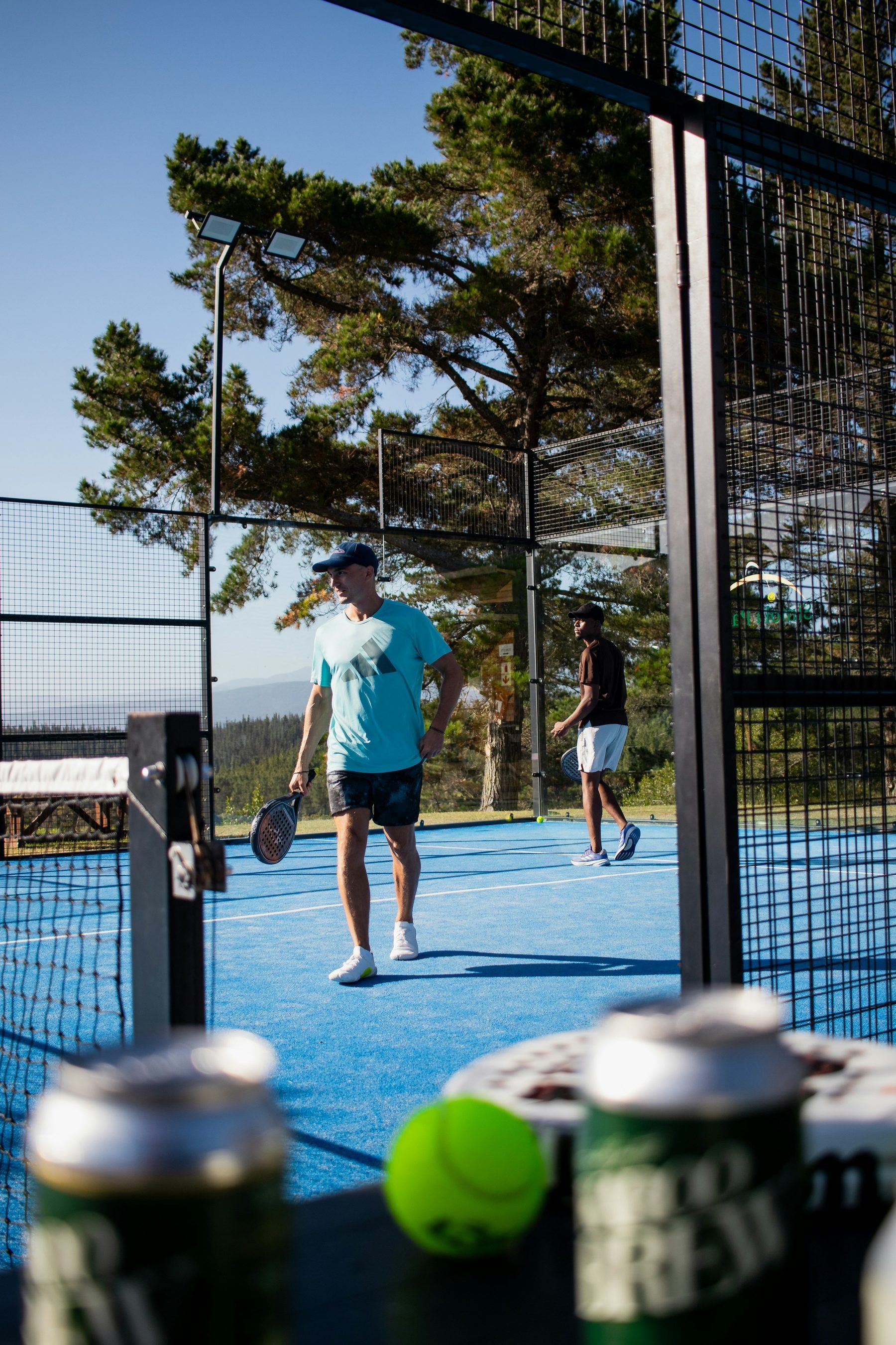Zwei Padelspieler auf einem Outdoor-Court zwischen Pinien, einer spielt den Ball von hinten