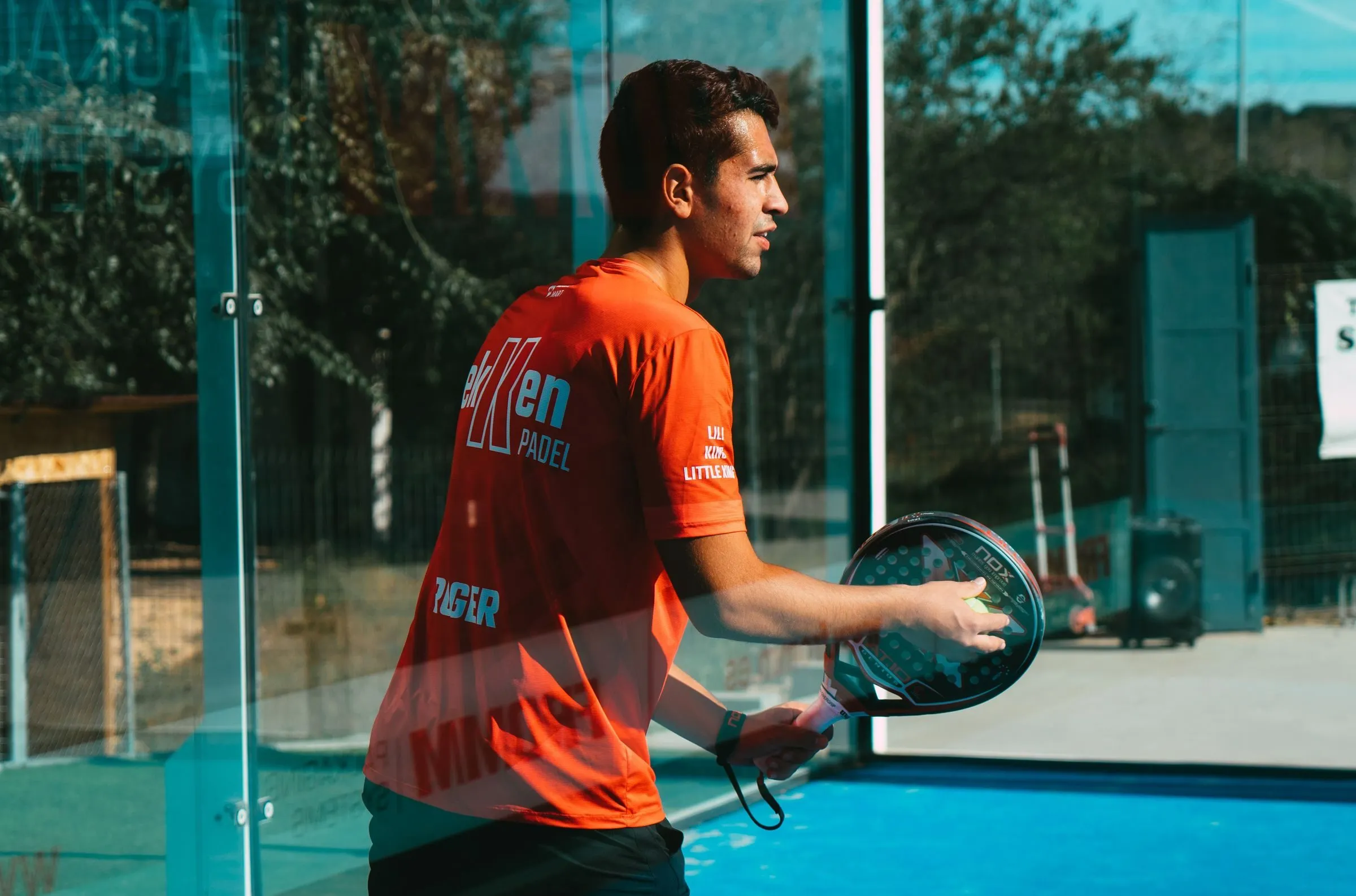 Padelspieler im orangefarbenen Shirt wartet hinter einer Glaswand auf einem Outdoor-Court auf den Ball