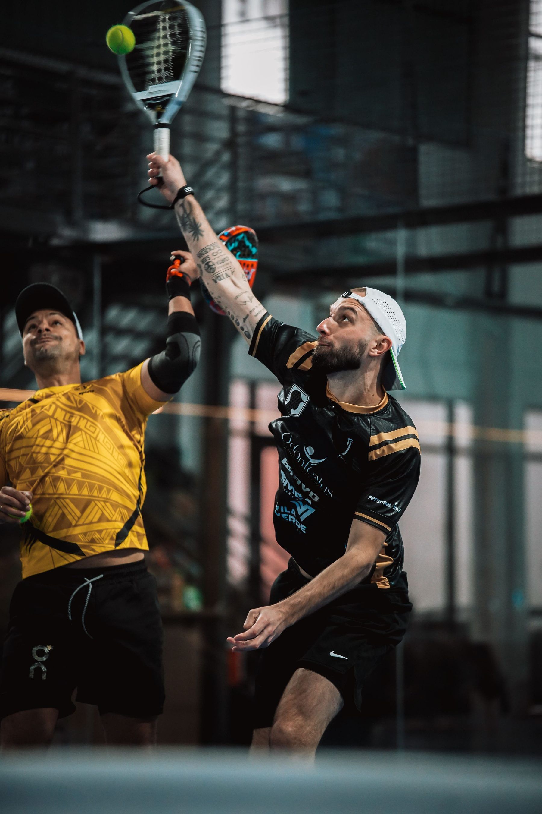 Zwei Padelspieler am Netz einer Halle, einer schlägt einen hohen Ball über Kopf