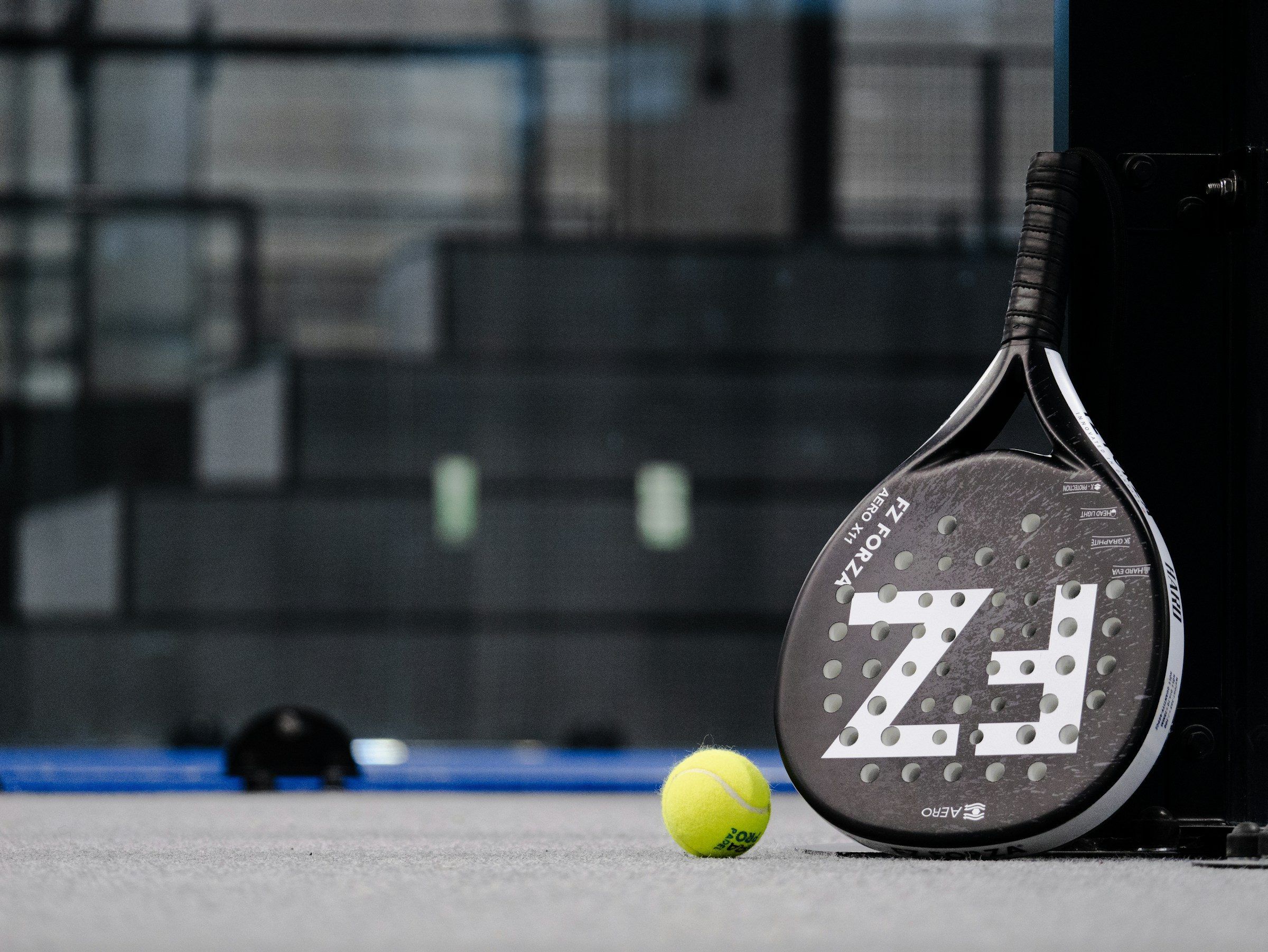 Padelschläger lehnt an der Wand, davor liegt ein gelber Ball auf dem Court