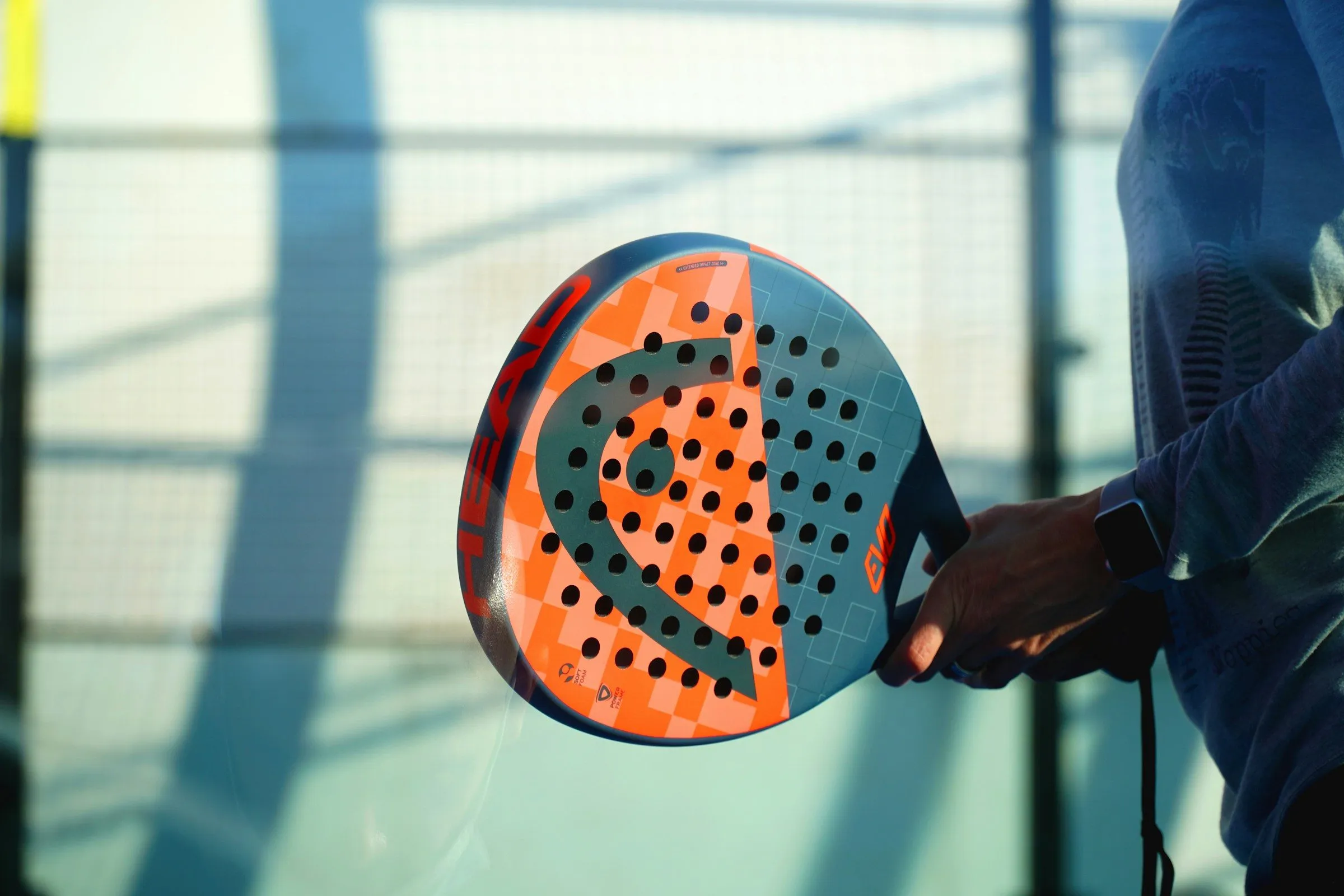 Hand hält einen orange-blauen Padelschläger mit gelochter Schlagfläche vor einer Glaswand