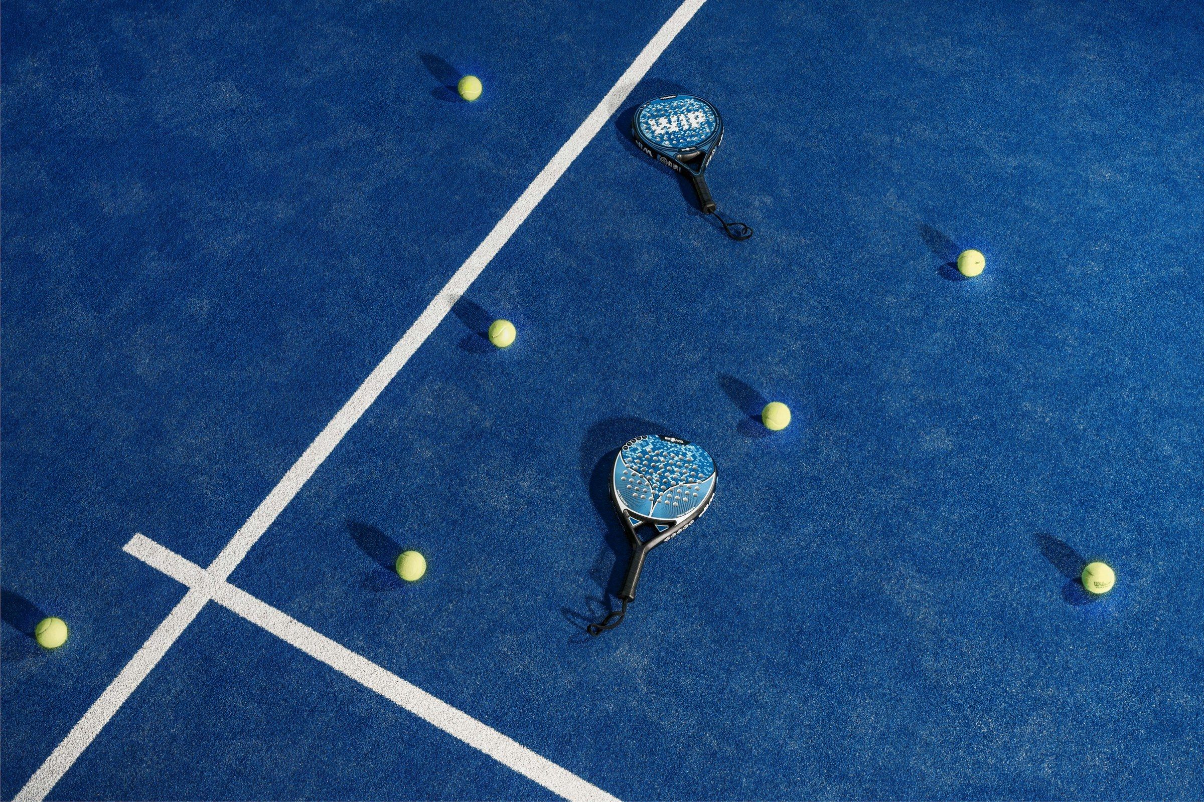 Zwei Padelschläger mit unterschiedlicher Kopfform liegen zwischen gelben Bällen auf einem blauen Court