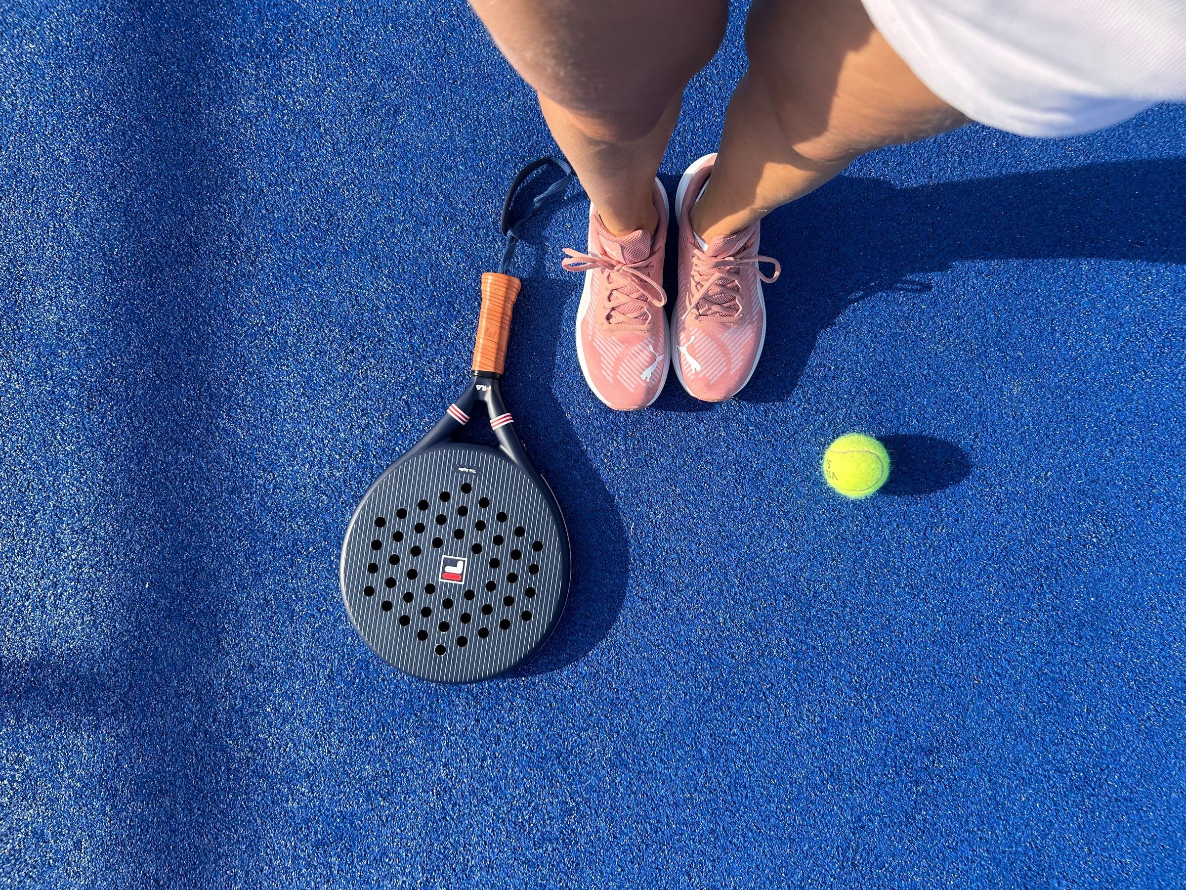 Füße in Sportschuhen neben einem Padelschläger und einem gelben Ball auf blauem Kunstrasen, von oben fotografiert
