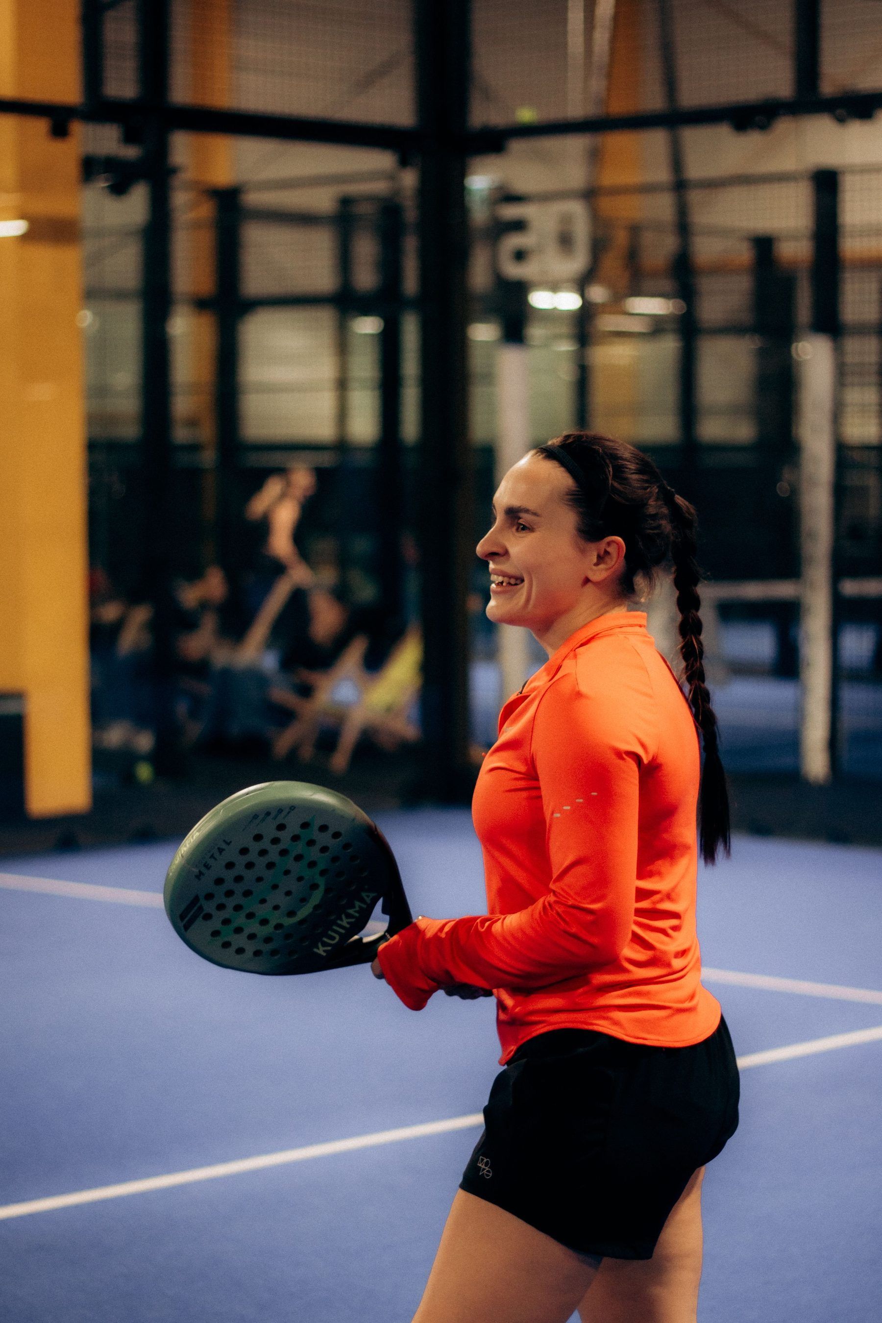 Padelspielerin in orangefarbenem Shirt hält den Schläger bereit auf einem Indoor-Court